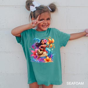 Puede incluir: Camiseta de color verde azulado con una ilustraci&oacute;n vibrante de un personaje tocando el ukelele, rodeado de flores coloridas y salpicaduras de agua. La palabra "SEAFOAM" est&aacute; impresa en la parte inferior. La camiseta la lleva una persona joven.