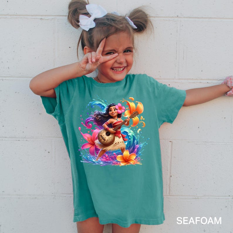 Puede incluir: Camiseta de color verde azulado con una ilustraci&oacute;n vibrante de un personaje tocando el ukelele, rodeado de flores coloridas y salpicaduras de agua. La palabra "SEAFOAM" est&aacute; impresa en la parte inferior. La camiseta la lleva una persona joven.