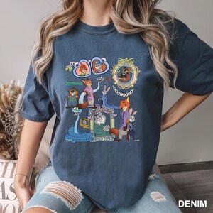 May include: Denim blue t-shirt featuring a colorful graphic of characters from the animated movie Zootopia. The design includes framed portraits and various animal characters. The word "Zootopia" is also visible on the shirt.