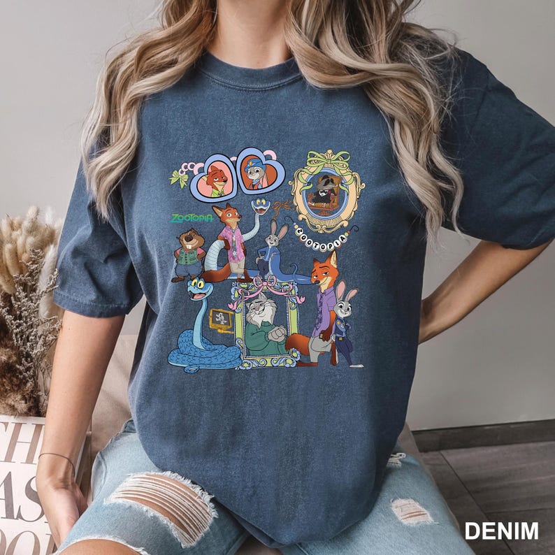 May include: Denim blue t-shirt featuring a colorful graphic of characters from the animated movie Zootopia. The design includes framed portraits and various animal characters. The word "Zootopia" is also visible on the shirt.