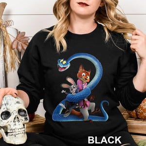 May include: Black sweatshirt featuring a cartoon illustration of characters from Zootopia, including Judy Hopps, Nick Wilde, and a blue snake. The word "BLACK" is printed at the bottom. The design is centered on the front of the sweatshirt.