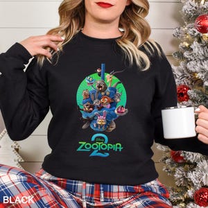 May include: Black crewneck sweatshirt featuring a colorful graphic of characters from the animated film "Zootopia 2". The design includes a green circle with various animal characters and the film's title in teal. The word "BLACK" is printed at the bottom.