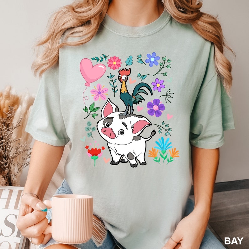 K&ouml;nnte beinhalten: Ein hellgr&uuml;nes T-Shirt mit einem Cartoon-Motiv aus einem Schwein und einem Hahn. Das Schwein ist wei&szlig; mit schwarzen Flecken und der Hahn ist rot, gelb und schwarz. Das Motiv ist von bunten Blumen und Bl&auml;ttern umgeben.