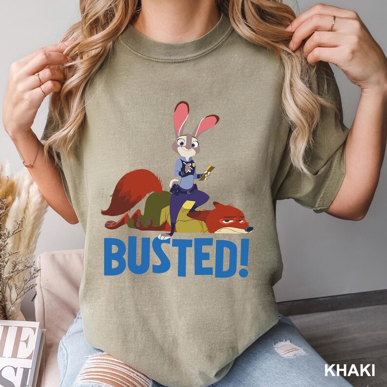 May include: Khaki-colored t-shirt featuring a cartoon image of Judy Hopps, a rabbit police officer, standing on Nick Wilde, a fox, with the word "BUSTED!" in blue. The shirt is made of a soft material.