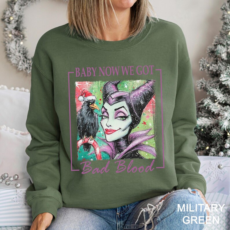 May include: Military green sweatshirt featuring a graphic of a cartoon villain with a raven wearing a Santa hat. The text "Baby Now We Got Bad Blood" is printed in a pink rectangle.