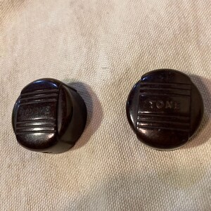 May include: Two dark brown, circular control knobs featuring a raised horizontal ribbed design on the top surface. Each knob has the word TONE embossed in capital letters across the center, surrounded by a smooth, rounded outer edge.