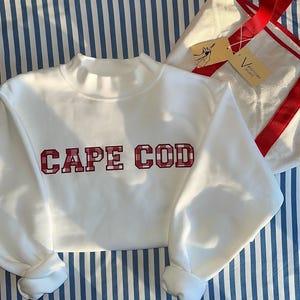 Op de afbeelding: Een witte sweatshirt met de woorden "CAPE COD" in rode geruite letters. De sweatshirt heeft een col en lange mouwen. Een witte draagtas met rode bandjes is zichtbaar op de achtergrond, rustend op een blauw-wit gestreept oppervlak.