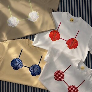 Puede incluir: Cuatro camisetas de manga corta en beige, blanco, amarillo claro y blanco. Cada camiseta presenta un estampado gráfico de una parte superior de bikini hecha con dos conchas marinas. Los diseños varían en color: blanco, rojo, azul y cuadros rojos.