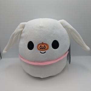 May include: A round white plush toy featuring long floppy ears, two black circular eyes, and an orange embroidered pumpkin nose. A thin pink band encircles the middle of the plush, and a small black smile is stitched below the pumpkin nose.