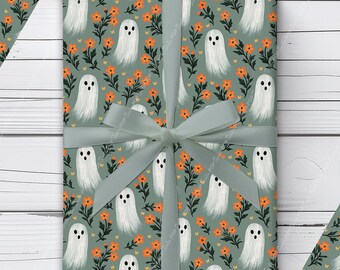 Puede incluir: Papel de regalo con un patrón repetitivo de fantasmas blancos y pequeñas flores naranjas sobre un fondo verde salvia. El papel está atado con una cinta verde salvia. El diseño incluye el texto Soeder POP en la esquina inferior izquierda.