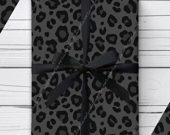Puede incluir: Un regalo rectangular envuelto en papel gris oscuro con un estampado de leopardo negro. El paquete está atado con una cinta de satén negra dispuesta en forma de cruz y rematado con un lazo centrado en la parte frontal del regalo.