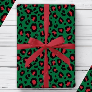 May include: A rectangular gift wrapped in green paper featuring a black and red leopard print pattern. The package is tied with a solid red ribbon that forms a bow in the center. The design includes a repeating animal spot motif throughout.