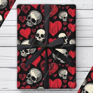 May include: A rectangular gift box wrapped in black paper featuring a repeating pattern of off-white human skulls and distressed red hearts. The package is tied with a black satin ribbon tied in a bow at the center, covering the surface.