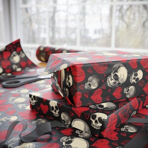 May include: Wrapping paper featuring a repeating pattern of off-white human skulls and red hearts on a black background. The paper is shown covering rectangular boxes, with a roll of the same paper and black scissors visible in the background.