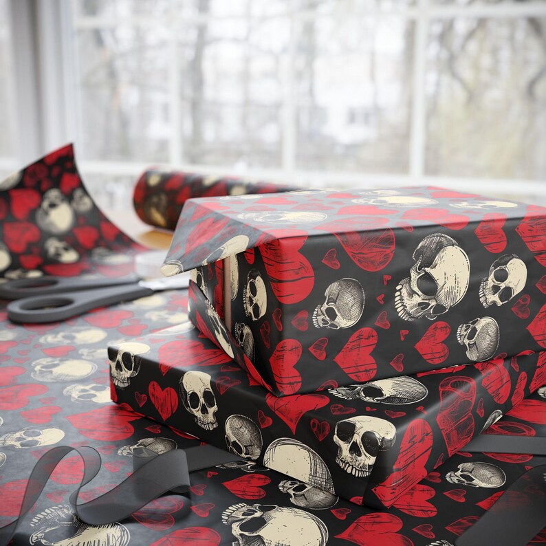 May include: Wrapping paper featuring a repeating pattern of off-white human skulls and red hearts on a black background. The paper is shown covering rectangular boxes, with a roll of the same paper and black scissors visible in the background.