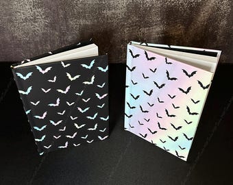 Puede incluir: Dos libros de tapa dura con páginas blancas. El libro de la izquierda tiene una cubierta negra con un patrón repetitivo de pequeños murciélagos iridiscentes de colores claros. El libro de la derecha tiene una cubierta iridiscente de colores claros con un patrón repetitivo de pequeños murciélagos negros.