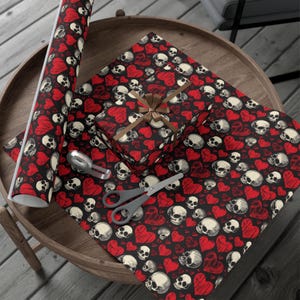 May include: A roll of wrapping paper and a gift box wrapped in the same paper, both featuring a repeating pattern of white skulls and red hearts on a black background. The gift box is tied with a brown ribbon, accompanied by scissors and tape.