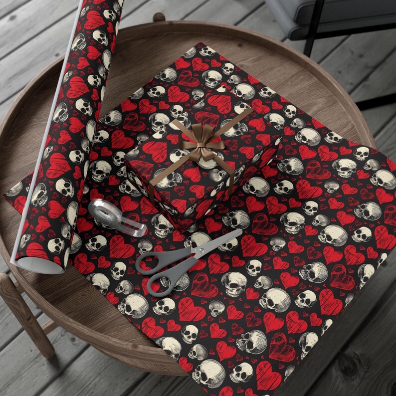May include: A roll of wrapping paper and a gift box wrapped in the same paper, both featuring a repeating pattern of white skulls and red hearts on a black background. The gift box is tied with a brown ribbon, accompanied by scissors and tape.