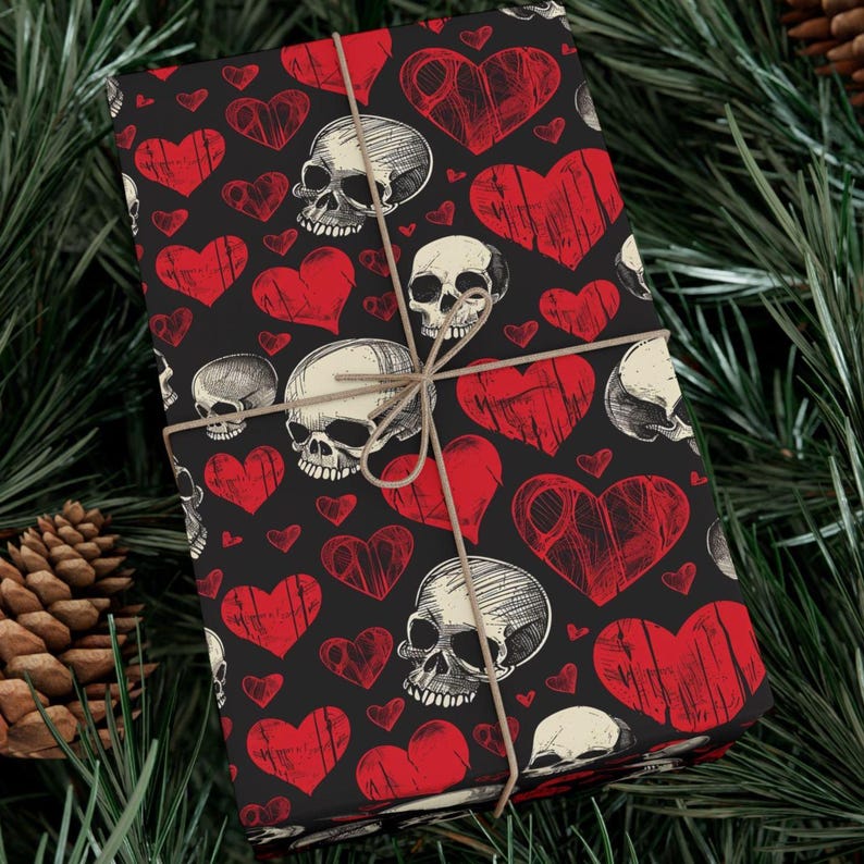 May include: A rectangular gift box wrapped in black paper featuring a repeating pattern of red hearts and white human skulls with detailed line shading. The package is tied with thin, light brown twine wrapped in a cross pattern around the center.