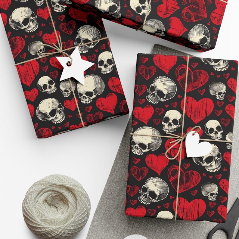 May include: Wrapping paper featuring a repeating pattern of off-white skulls and red hearts on a black background. The paper is tied with brown twine and includes a white star-shaped gift tag on one package and a heart-shaped tag on another.