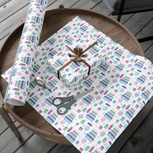 May include: White wrapping paper with a repeating pattern of colorful birthday cakes and balloons. The paper is rolled up and tied with a brown ribbon.