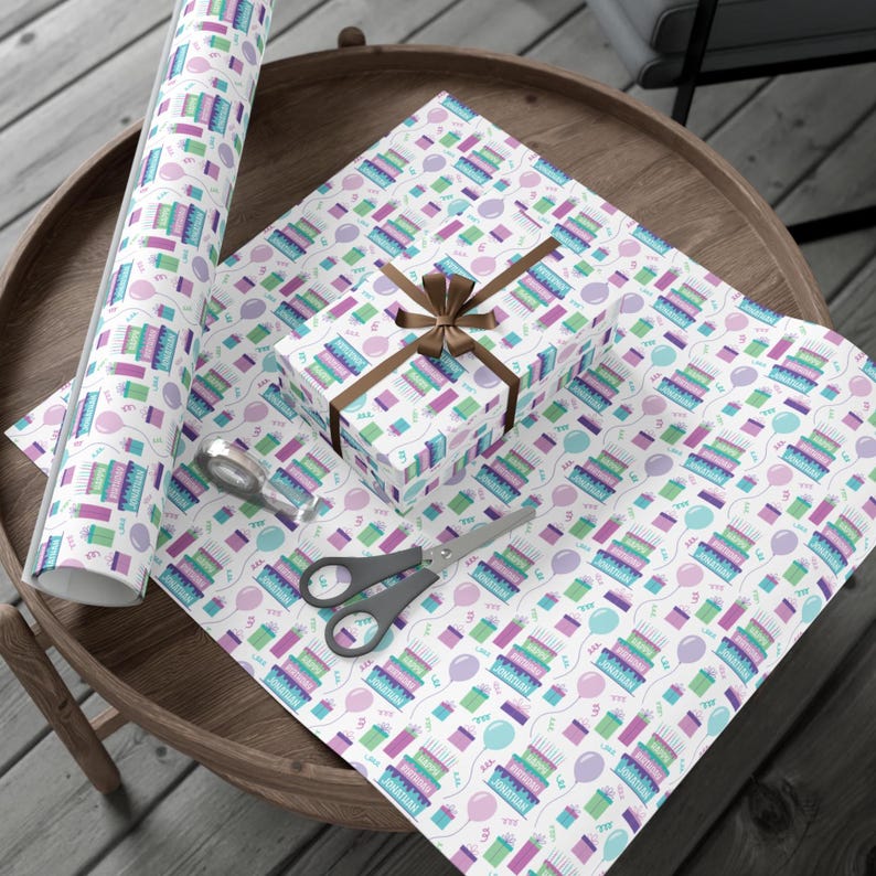 May include: White wrapping paper with a repeating pattern of colorful birthday cakes and balloons. The paper is rolled up and tied with a brown ribbon.