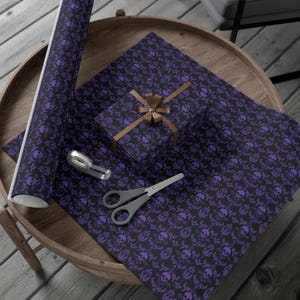 May include: Black wrapping paper with a purple skull and floral design, a wrapped gift box, scissors, and a tape dispenser on a wooden table. The gift box is tied with a brown ribbon. The items are arranged for gift wrapping.