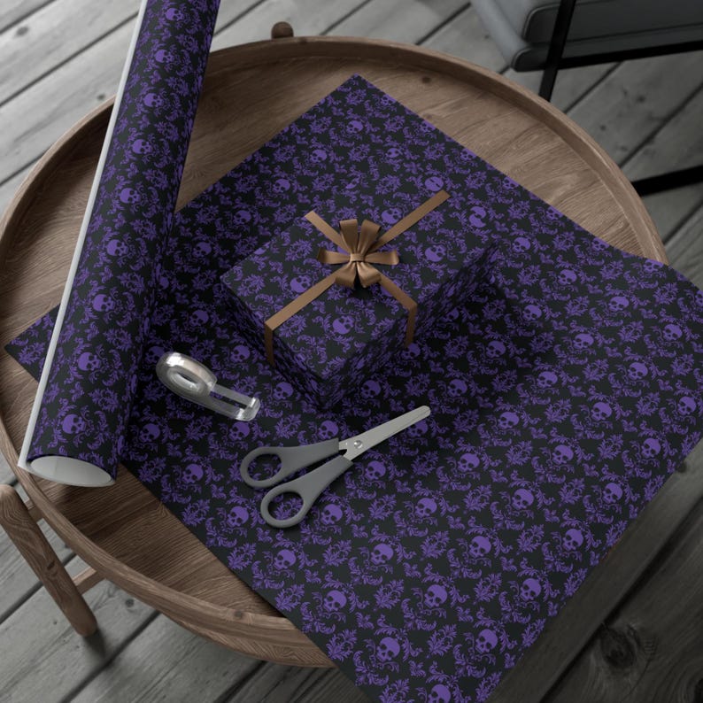 May include: Black wrapping paper with a purple skull and floral design, a wrapped gift box, scissors, and a tape dispenser on a wooden table. The gift box is tied with a brown ribbon. The items are arranged for gift wrapping.