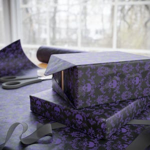 May include: Gift-wrapped boxes with black and purple skull and damask patterned paper. A roll of matching paper, scissors, and a grey ribbon are also visible, suggesting a Halloween or gothic theme.