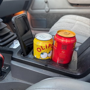 May include: A yellow can with a cartoon face and the word "OLIPOP" next to a red can in a car's cup holder. A black cell phone is in the phone holder. The interior of the car is gray.