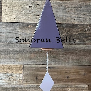 May include: A purple triangular wind chime with a wooden base and a diamond-shaped clapper. The text "Sonoran Bells" is written on the chime. The chime hangs from a chain against a rustic wooden background.