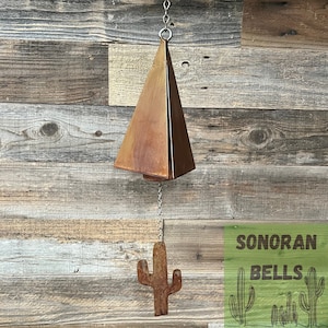 May include: A rusty metal wind chime shaped like a triangle with a cactus-shaped charm hanging from it. The wind chime is hanging from a chain against a wood background. The text "SONORAN BELLS" is visible in the lower right corner of the image.