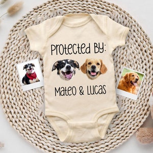 May include: A cream-colored baby onesie with the text "Protected By: Mateo & Lucas" and two dog photos printed on the front.