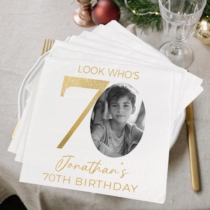 May include: White paper napkins with gold glitter text that reads "Look Who's 70 Jonathan's 70th Birthday". A photo of a boy is in the center of the napkin.