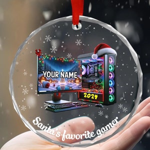 May include: A clear, round Christmas ornament with a red ribbon. The ornament features a computer with a Santa hat, a monitor displaying a winter scene, and the text "Your Name" and "2024". The text "Santa's favorite gamer" is also visible.