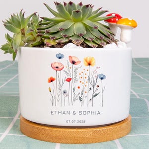 May include: A white ceramic planter with a floral design, holding a green succulent plant. The planter features the names "ETHAN & SOPHIA" and the date "07.07.2025". Two decorative mushroom figurines sit atop the plant, and the planter rests on a wooden base.