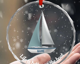 May include: A clear glass Christmas bauble featuring a sailboat design and the text "Life is better on the boat". The bauble has a red ribbon attached to the top.