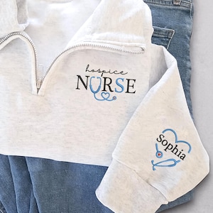 May include: A light gray quarter-zip sweatshirt with the words "hospice NURSE" and a stethoscope design. The sleeve features a heart-shaped stethoscope and the name "Sophia". The sweatshirt is paired with blue jeans.