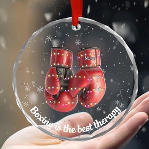 May include: A clear glass Christmas ornament with a red ribbon. The ornament features a pair of red boxing gloves with white lights around them. The text "Boxing is the best therapy" is written on the ornament.