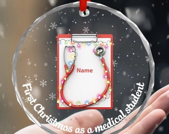 May include: A clear glass Christmas ornament with a red ribbon. The ornament features a red stethoscope with colorful lights wrapped around it, a white clipboard with the word "Name" written on it, and the text "First Christmas as a medical student".