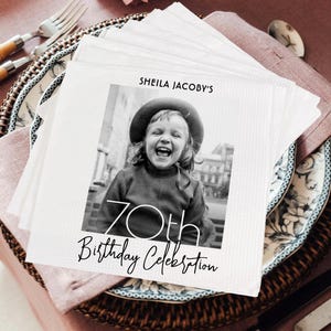 May include: A white napkin with black text that reads "Sheila Jacoby's 70th Birthday Celebration". A black and white photo of a young girl wearing a hat is in the center of the napkin.