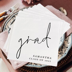May include: White paper napkin with the word "grad" written in black cursive script. Below the word is the text "Samantha Class of 2025".