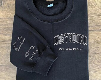 Peut inclure: Sweat-shirt noir avec le texte "Greyhound Mom" en blanc sur le devant. Les manches portent les noms "Charlie" et "Allie" en blanc.