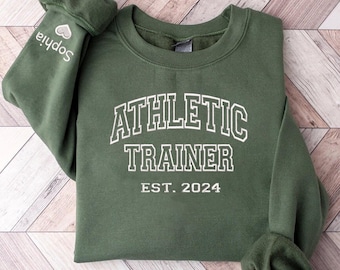 May include: A dark green sweatshirt with the text "ATHLETIC TRAINER EST. 2024" in white outlined letters.