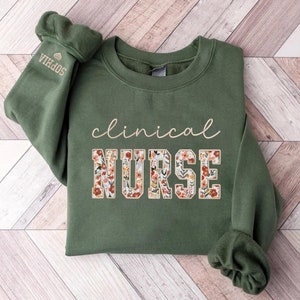 May include: A green sweatshirt with the text "clinical nurse" in white lettering. The word "nurse" is decorated with a floral pattern in shades of orange, pink, and yellow.