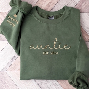 May include: A green sweatshirt with the word "auntie" in gold script and the text "EST. 2024" below it. The sweatshirt has a small heart on the left sleeve with the text "Brian Sophia" above it.