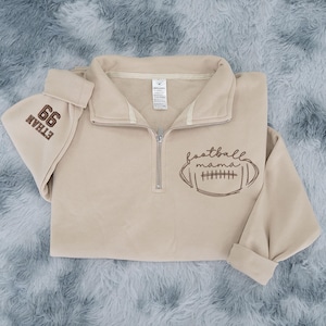 May include: A beige quarter-zip sweatshirt featuring a brown embroidered football graphic with the text "football mama" on the front. The sleeve is embroidered with the number "99" and the name "ETHAN" in brown thread. It has a silver zipper.