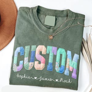 May include: Olive green t-shirt with the word "CUSTOM" in patchwork letters. Below the word are the names "Sophia James Noah". A gold necklace with a pendant is draped across the shirt.