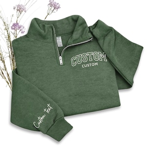 May include: A green quarter-zip sweatshirt with the word "CUSTOM" in white block letters on the chest and the words "Custom Text" on the sleeve. The sweatshirt has a silver zipper and is folded on a white surface.