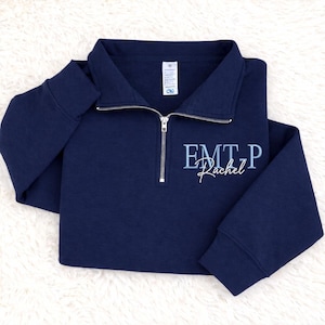 May include: A navy blue quarter-zip sweatshirt featuring a silver-toned metal zip and a fold-over collar. The front is embroidered with the light blue text EMT-P above the white cursive name Rachel, positioned on the right side of the chest.